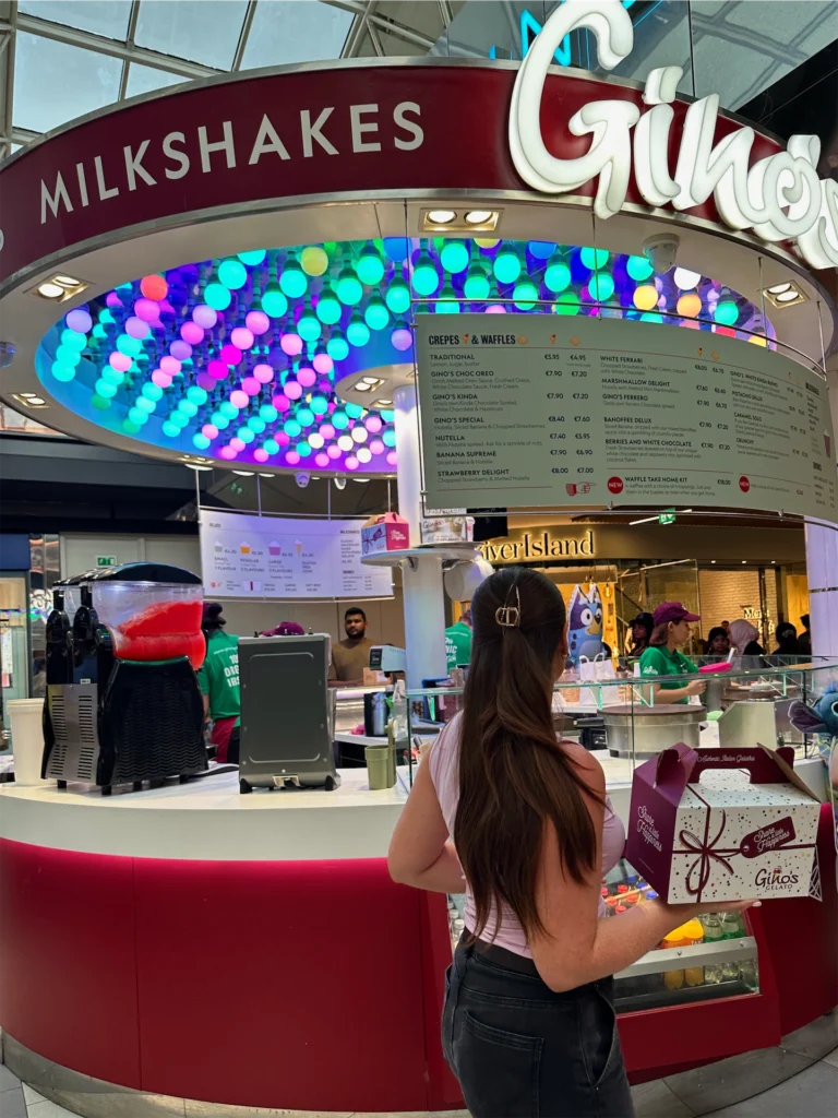 Customer at colourful milkshake stall in Dublin shopping centre, holding a Gino's box. Menu and staff visible.